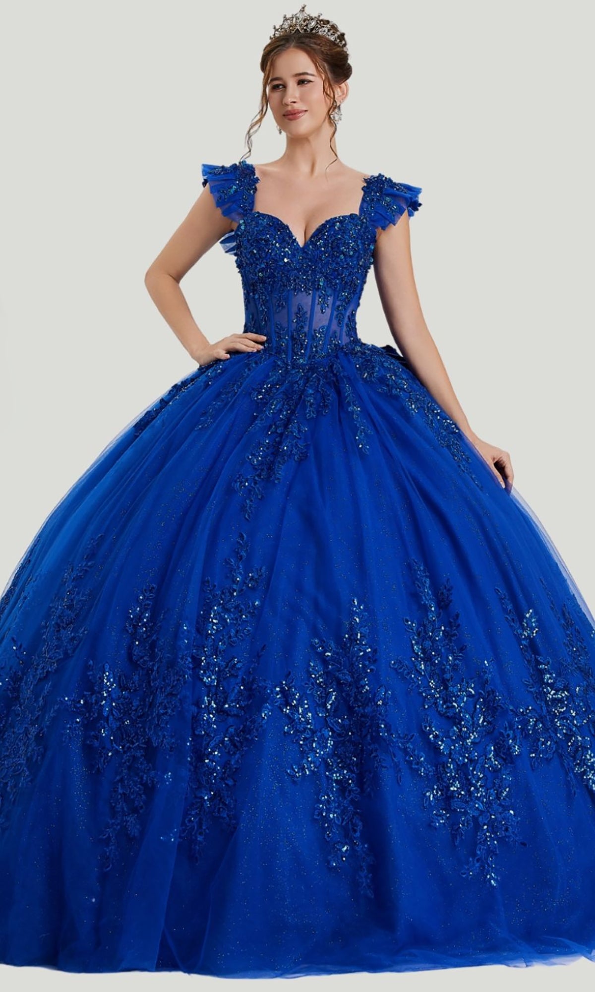 A woman wearing the Quinceanera Dress 26410 By Stella Couture in royal, featuring shimmering sequin embellishments, an embellished bodice, off-shoulder sleeves, and a voluminous tulle skirt, stands against a plain background.